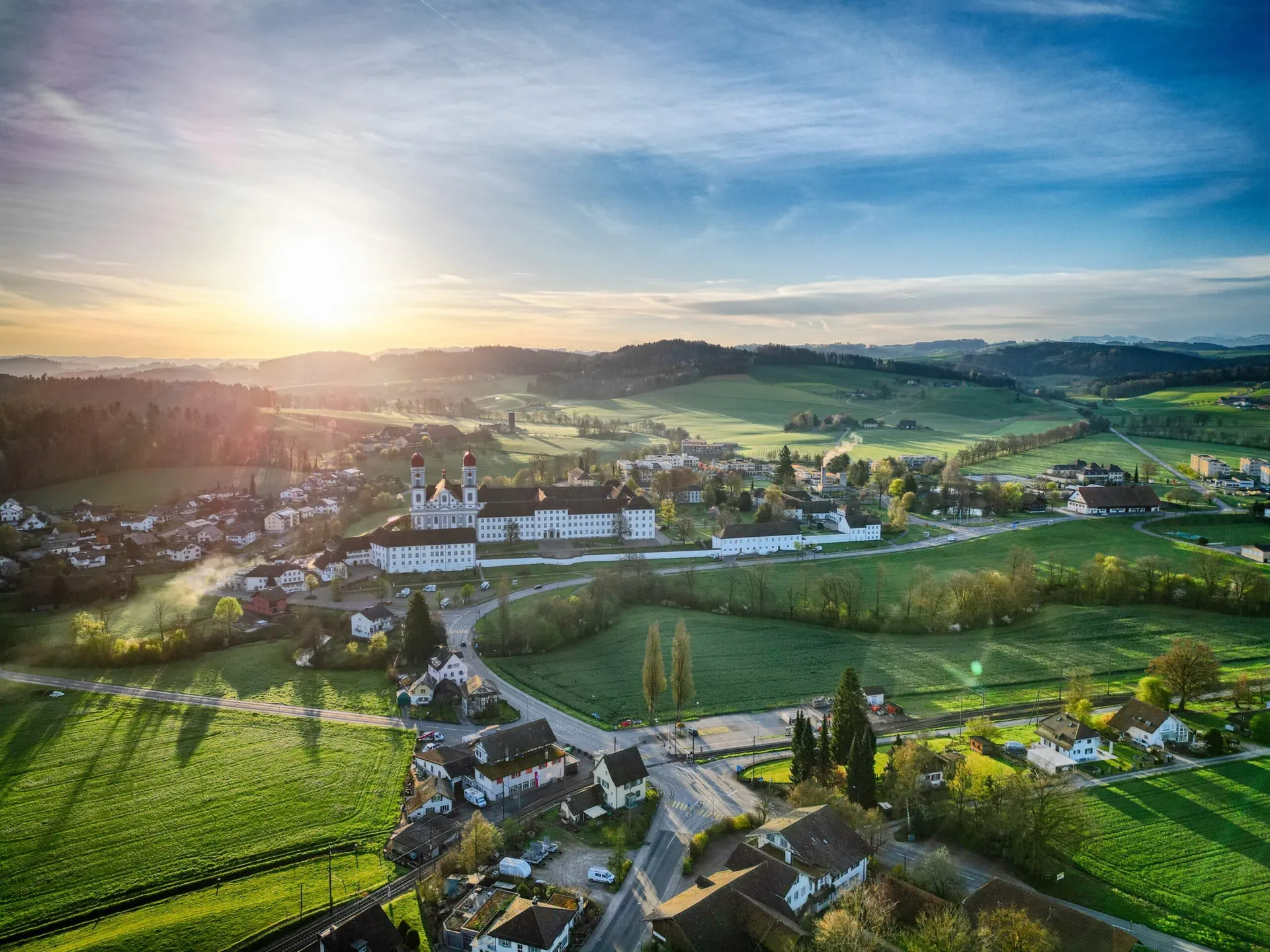 Drohnenaufnahme von Pfaffnau bei Sonnenaufgang: Das Dorf liegt zwischen grünen Feldern und Hügeln, die Sonne geht über dem Horizont auf.