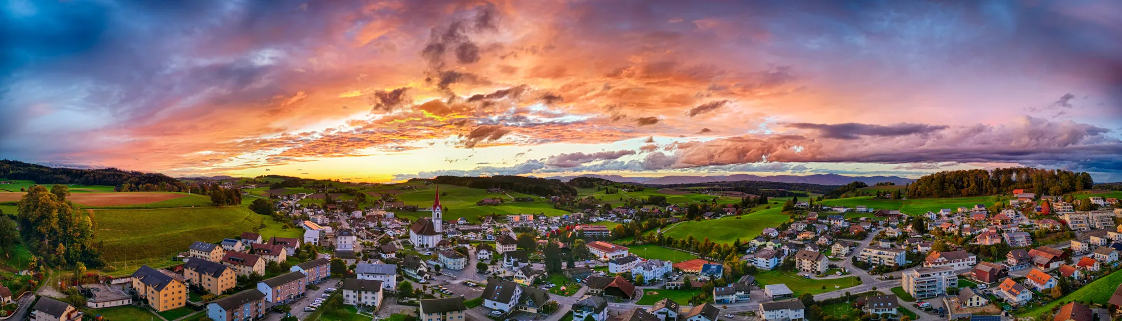 Weites Abendpanorama über Pfaffnau: Der Kirchturm steht mitten im Dorf, der Himmel glüht orange und violett über den Hügeln.