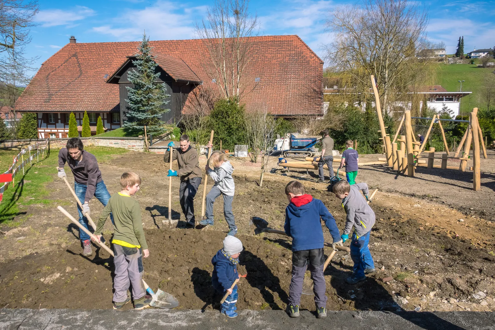 Familien aus dem Dorf bauen mit Schaufeln und Pickeln am Generationenpark. Kinder und Erwachsene arbeiten zusammen, im Hintergrund steht ein Bauernhaus.
